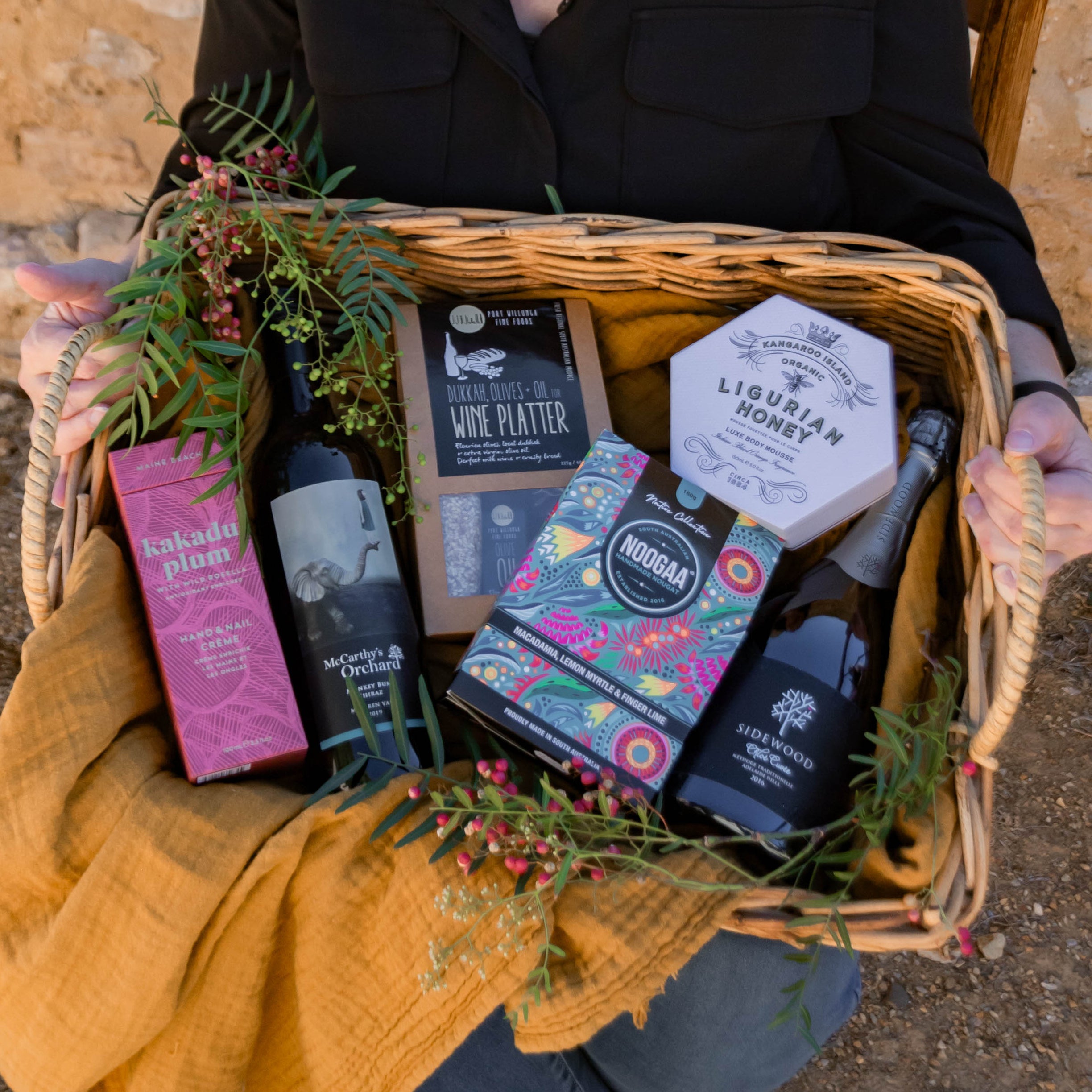 A woman wearing glasses and a black shirt sits outdoors against a rustic stone wall, holding a wicker gift basket lined with a mustard yellow blanket and decorated with greenery. The basket contains assorted packaged goods, including a pink box, a dark bottle, a white box, and a colorful patterned package.