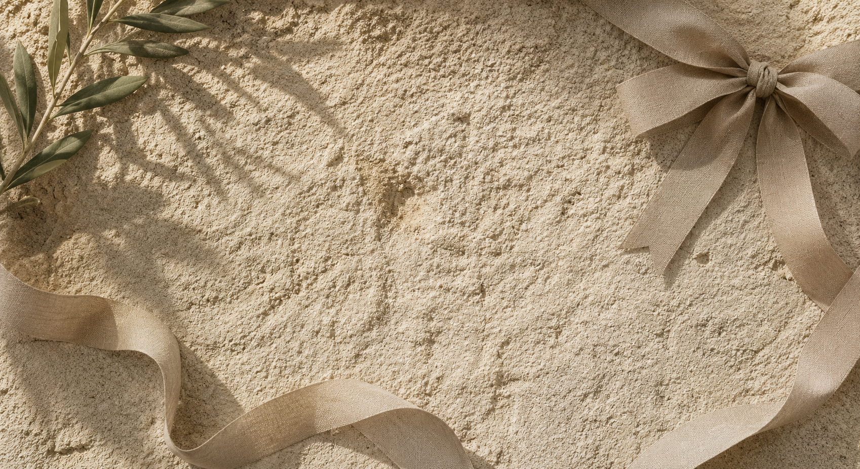 A styled flat-lay featuring a textured beige stone surface, a beige satin ribbon bow in the upper right, a beige ribbon flowing across the lower left, and a green olive branch in the upper left with leaf shadows cast across the stone.
