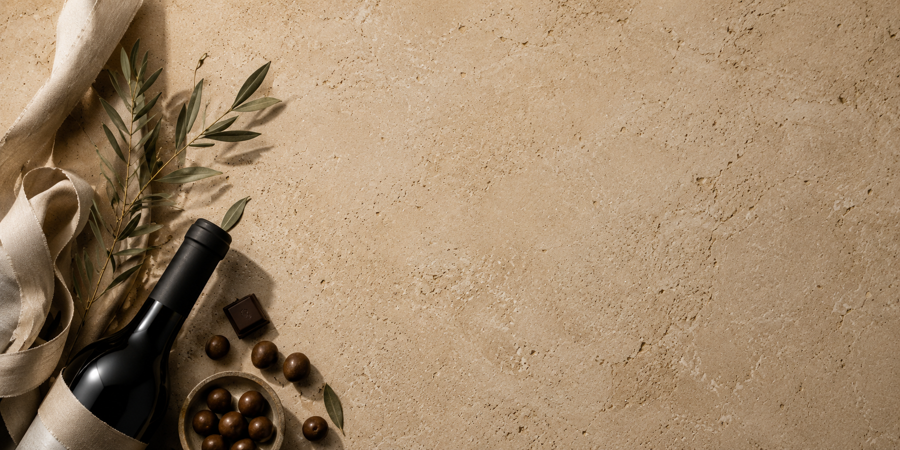 A dark wine bottle with a beige fabric sleeve, styled with olive branches, olives, and chocolate pieces on a textured beige stone surface with negative space on the right.