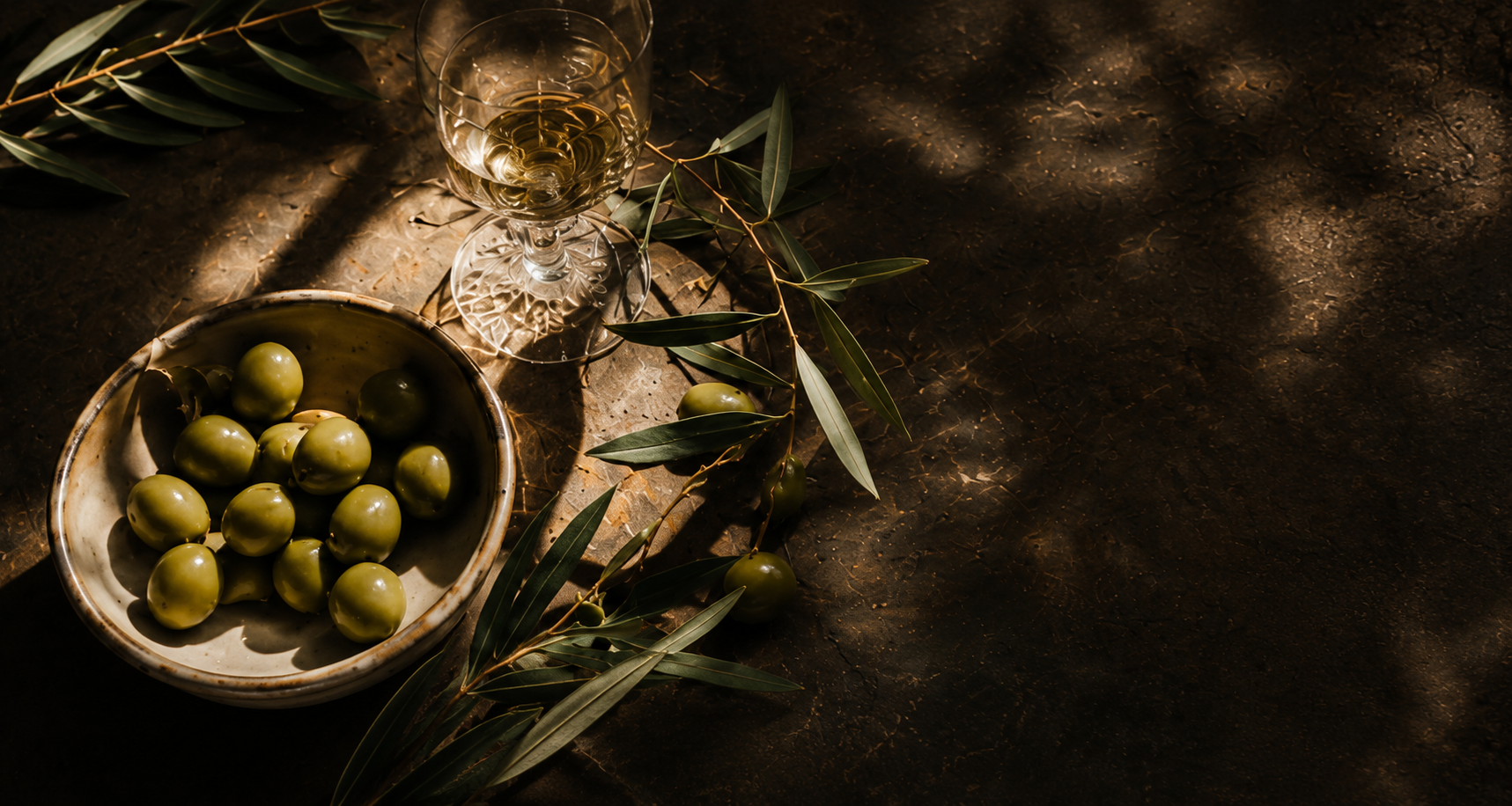 A rustic bowl of green olives and olive branches arranged on a dark stone surface next to a glass of white wine, with dramatic dappled sunlight and shadows.